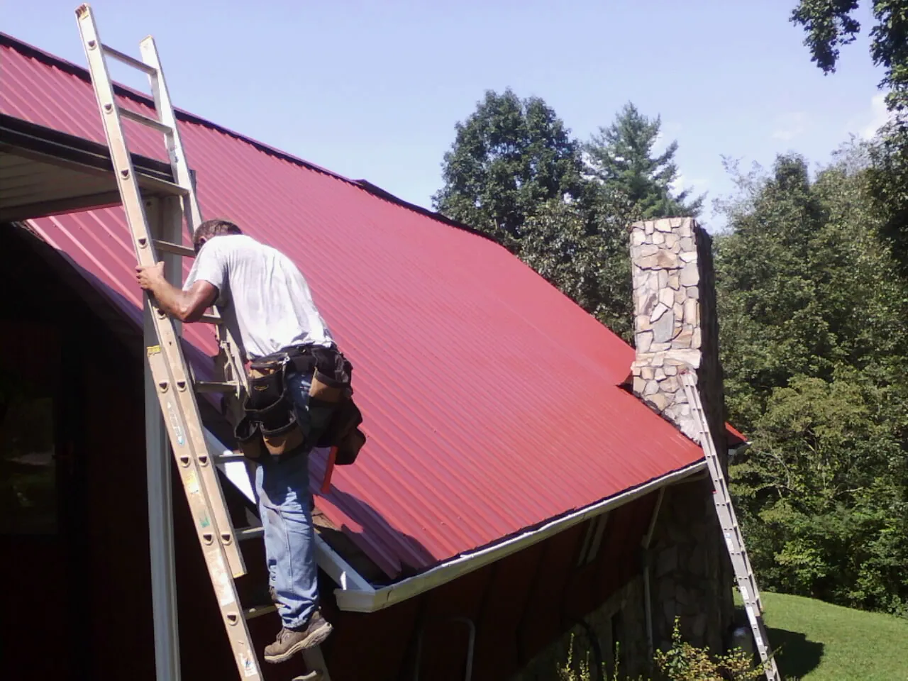 Roofer on a ladder beside a red metal roof and stone chimney — Soffit & Fascia Repair by Roofing Brothers in Ridgway